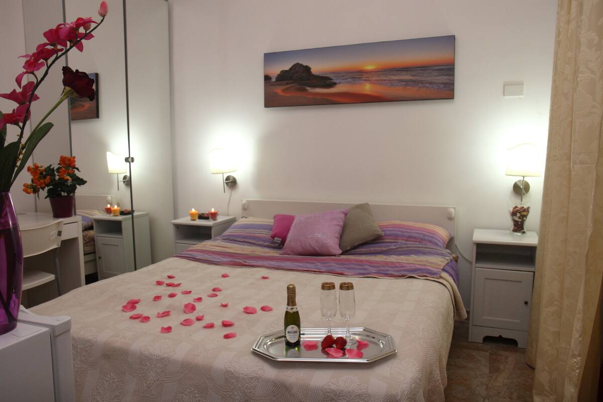 A hotel room with a bed, bedside tables, a painting, and a tray with rose petals and champagne.