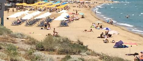 Una playa con filas de tumbonas y sombrillas, un cielo despejado y montañas al fondo.