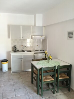 A compact kitchen with white cabinets, a stove, and a sink. There is a dining table with chairs and a vase with flowers. - Stella Apartments (Hersonissos)