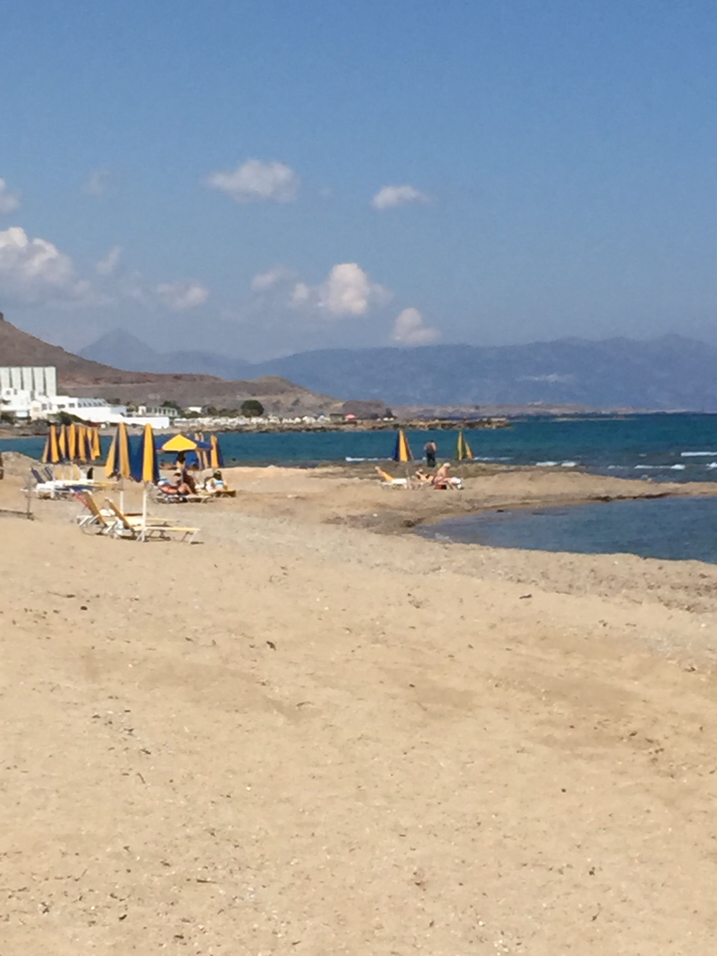Una playa con tumbonas y sombrillas, cielo despejado y montañas a lo lejos.