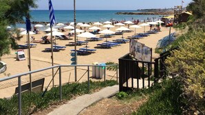 A beach with rows of sun loungers and umbrellas, a flagpole with a flag, and a lifeguard tower. - Stella Apartments (Hersonissos)