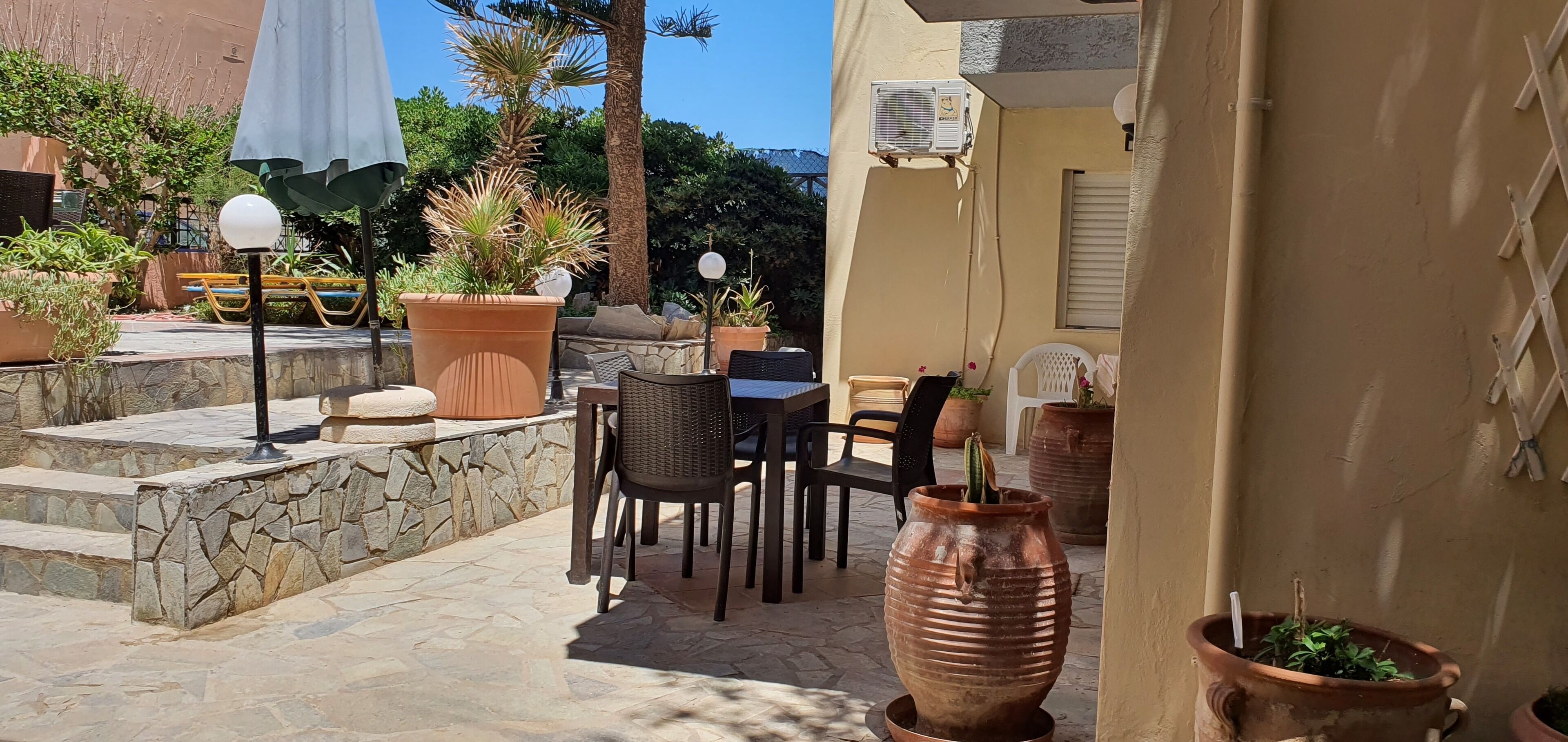 Un patio al aire libre con escalones de piedra, una mesa con sillas, plantas en macetas y una maceta de terracota grande.