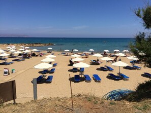 A beach with rows of sun loungers and umbrellas, a rocky shoreline, and clear blue water. - Stella Apartments (Hersonissos)