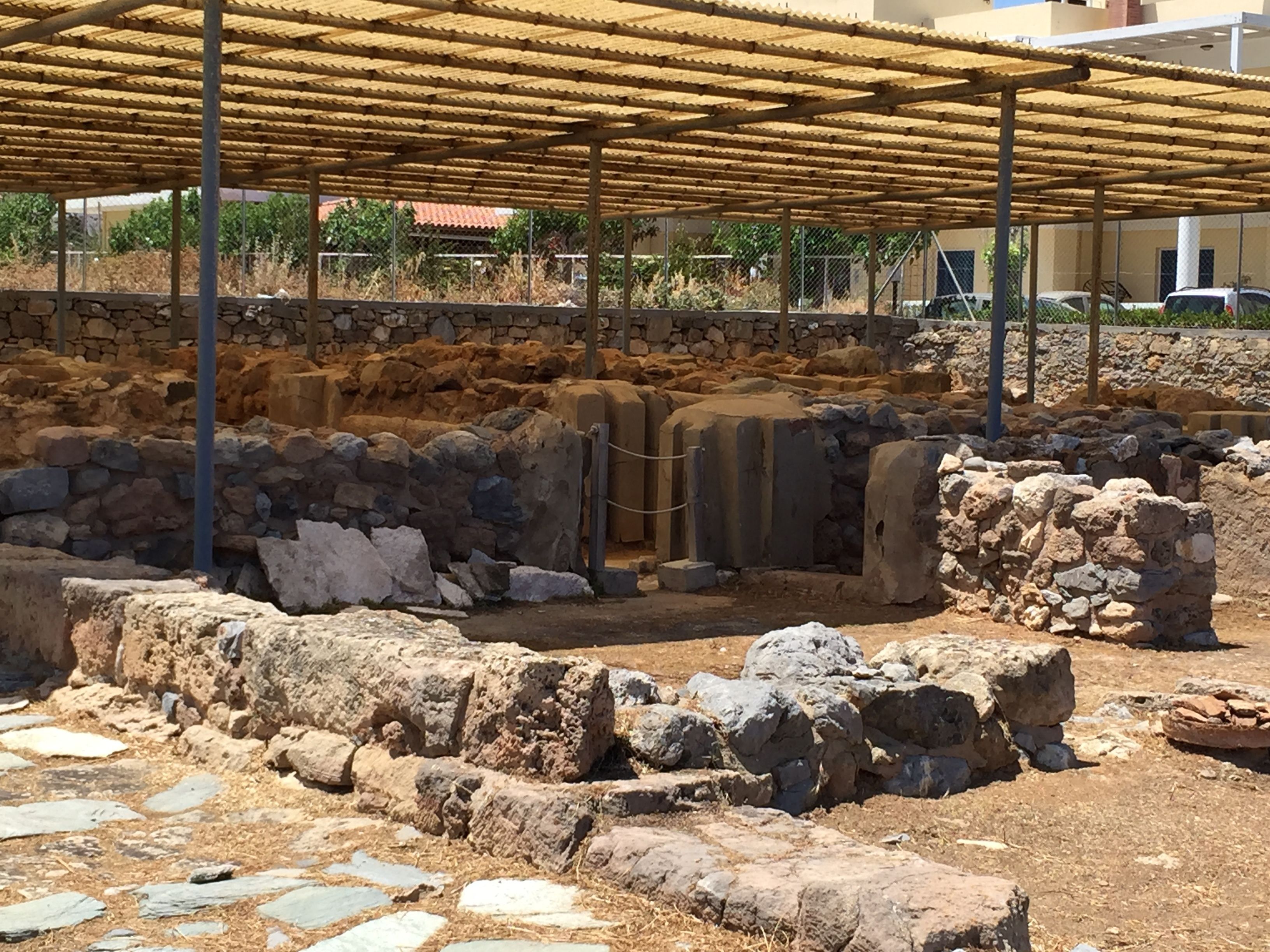 Sítio arqueológico com estruturas de pedra e cobertura metálica.