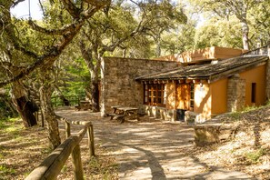 Un bâtiment rustique en pierre, agrémenté d’une table et de bancs en bois pour pique-niquer, entouré d’arbres et bordé d’un sentier en pierre.