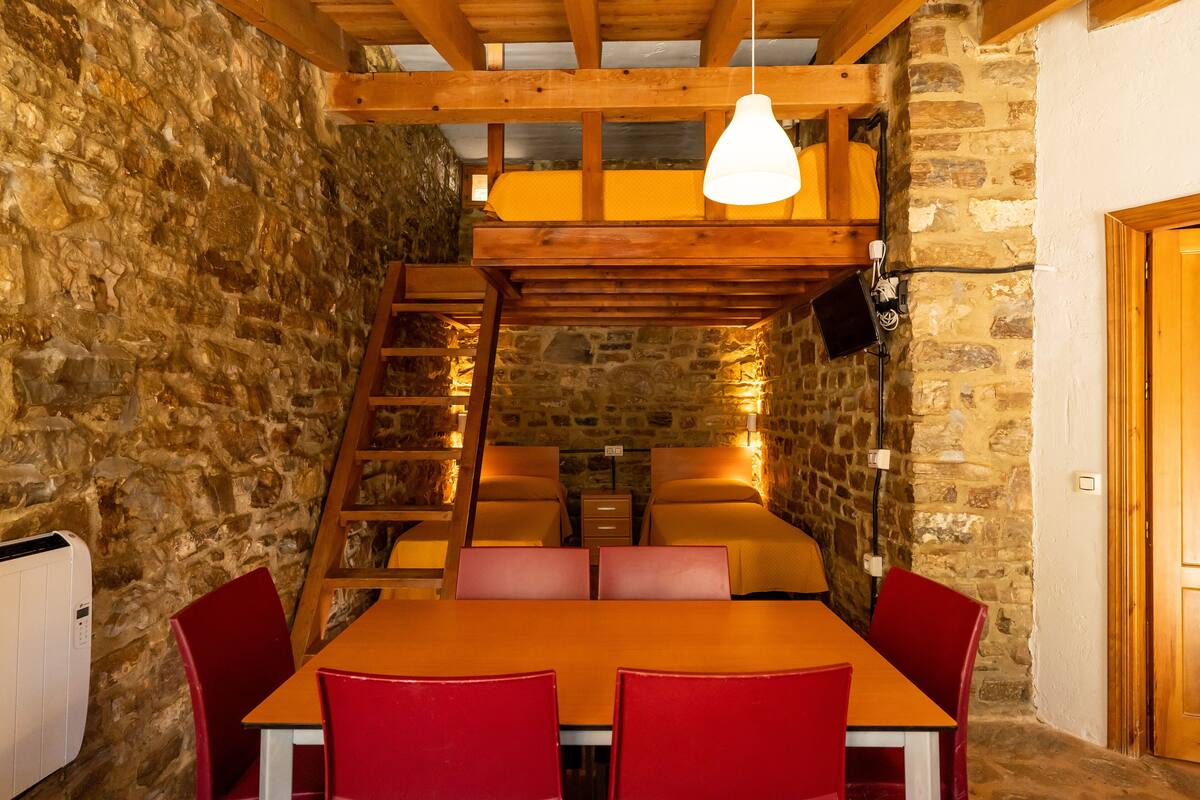Una habitación rústica con pared de piedra, vigas de madera, cama en alto, mesa de comedor con sillas rojas y una lámpara colgante.