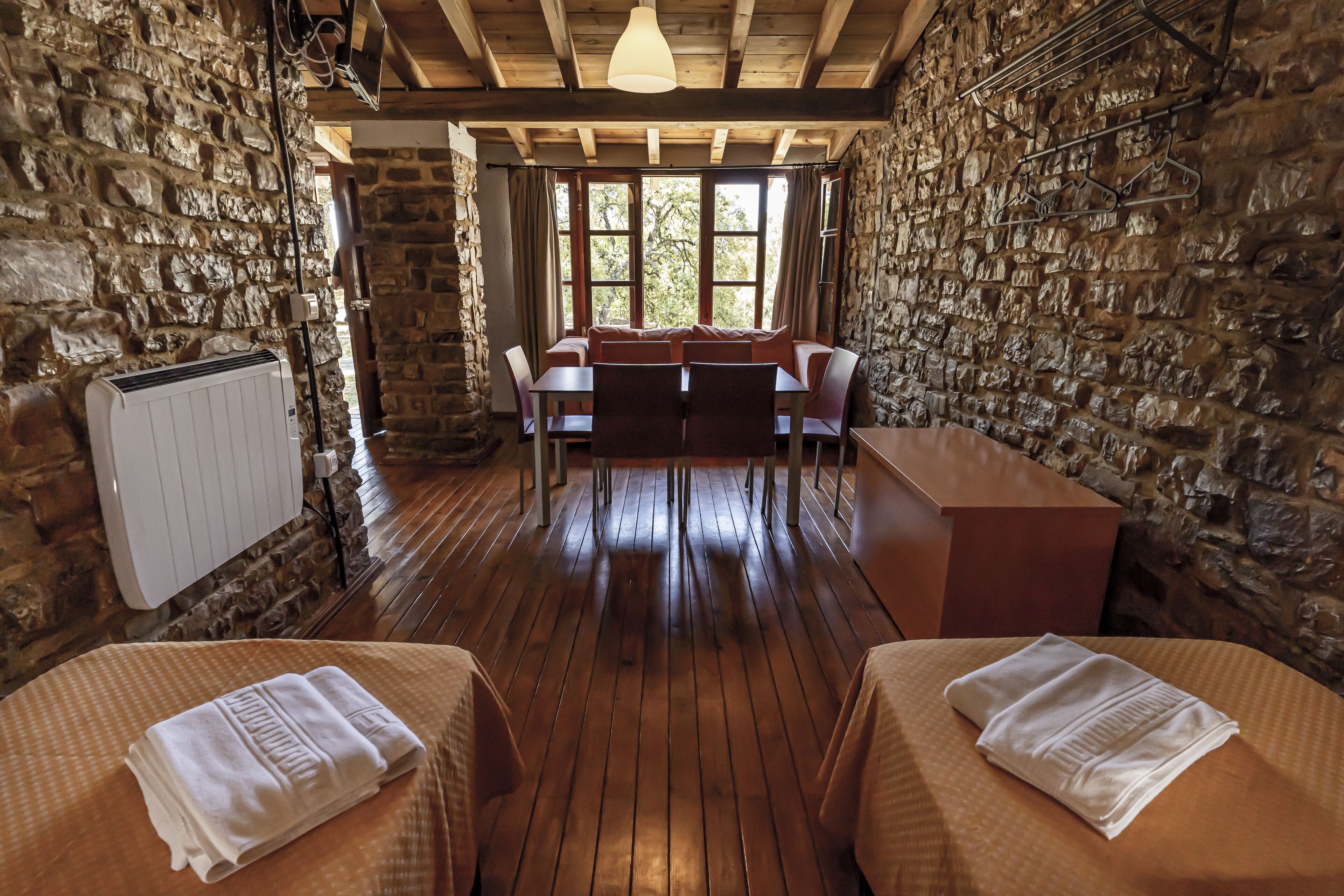 Una habitación rústica con paredes de piedra, piso de madera, una mesa de comedor con sillas y dos camas con toallas.
