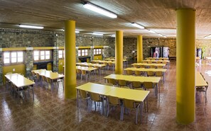 A spacious room with multiple yellow tables and chairs, stone walls, and a tiled floor. - Albergue Inturjoven Cortes de la Frontera - Hostel (Cortes de la Frontera)