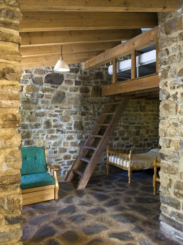 Une pièce rustique aux murs de pierre, avec une échelle en bois, un fauteuil recouvert d’un tissu vert et un lit en bois.