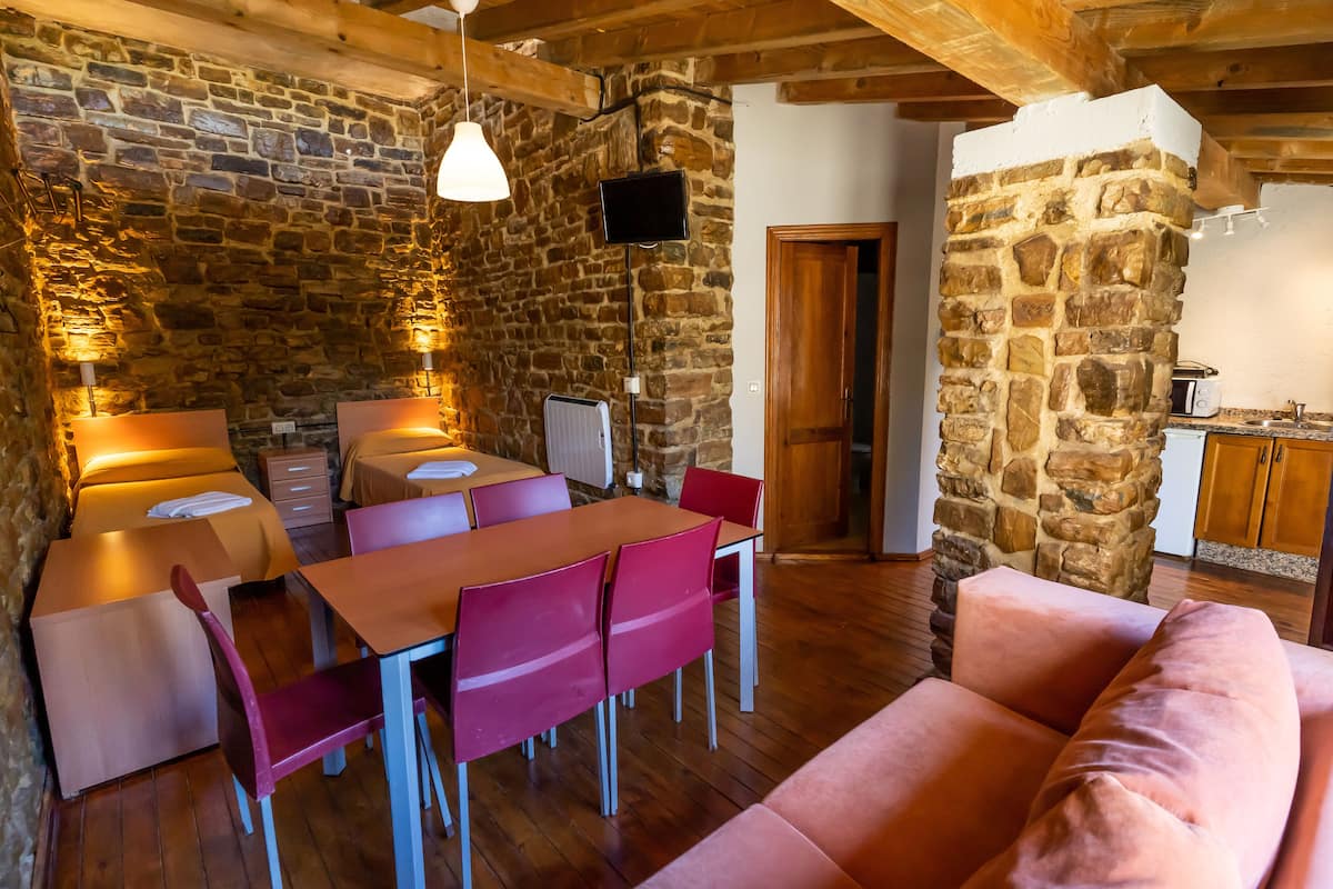 Un espacio acogedor con pared de piedra, vigas de madera en el techo, una mesa de comedor con sillas, un sofá rosa y una zona de cocina con un pilar de piedra.