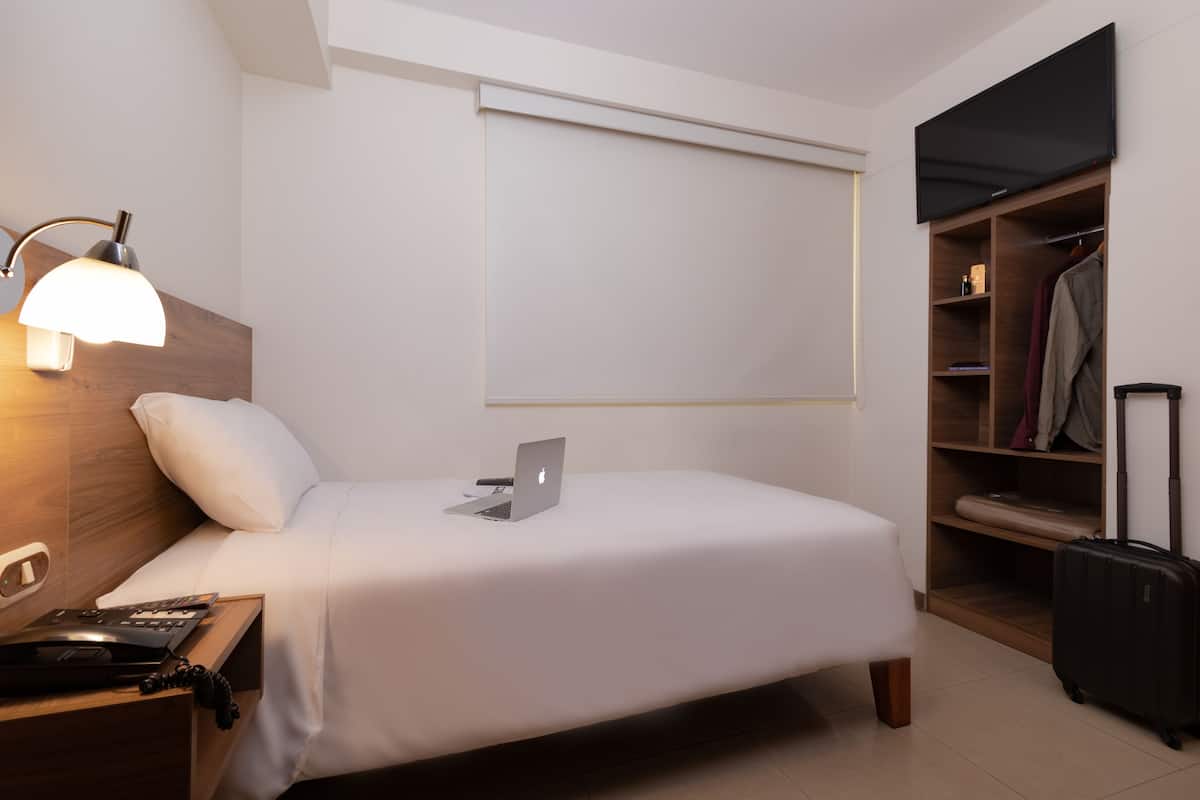 Habitación de hotel con cama, escritorio con lámpara, ordenador portátil, maleta y armario.
