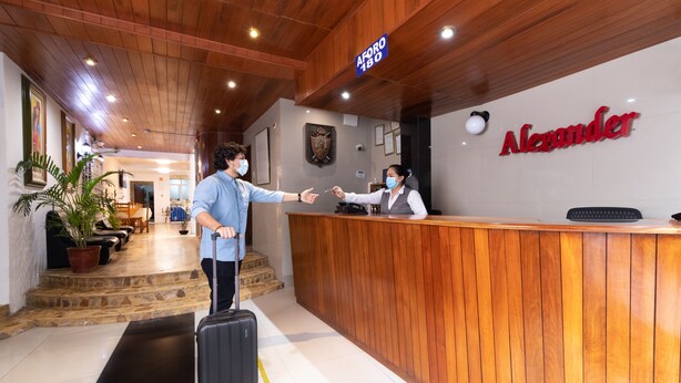 A hotel reception area with a wooden reception desk, a sign reading "AFORE 130", and a receptionist. - Hotel Alexander (Trujillo)