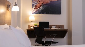 A hotel room with a bed, a desk with a laptop, a chair, a lamp, a picture of a coastal landscape, and a wall-mounted light fixture. - Hotel Alexander (Trujillo)