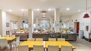 Un comedor amplio con mesas amarillas y sillas blancas, una cocina al fondo y una lámpara colgante roja.