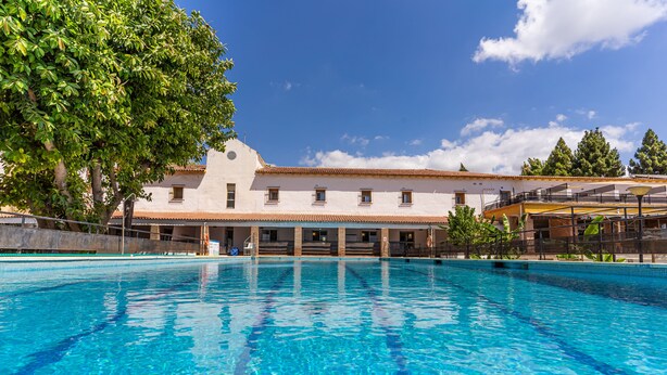 Una piscina con agua azul cristalina, un edificio blanco con techo de tejas y un árbol grande a la izquierda.