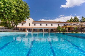 Una piscina con agua azul cristalina, un edificio blanco con techo de tejas y un árbol grande a la izquierda.