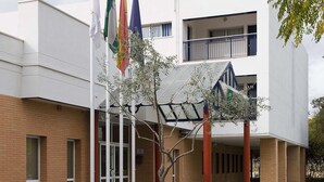 A building with a modern architectural style, featuring a flat roof and a two-story structure. There are multiple flags displayed prominently in front of the building, and a covered outdoor area with a tree in the center. - Albergue Inturjoven Sevilla - Hostel (Seville)