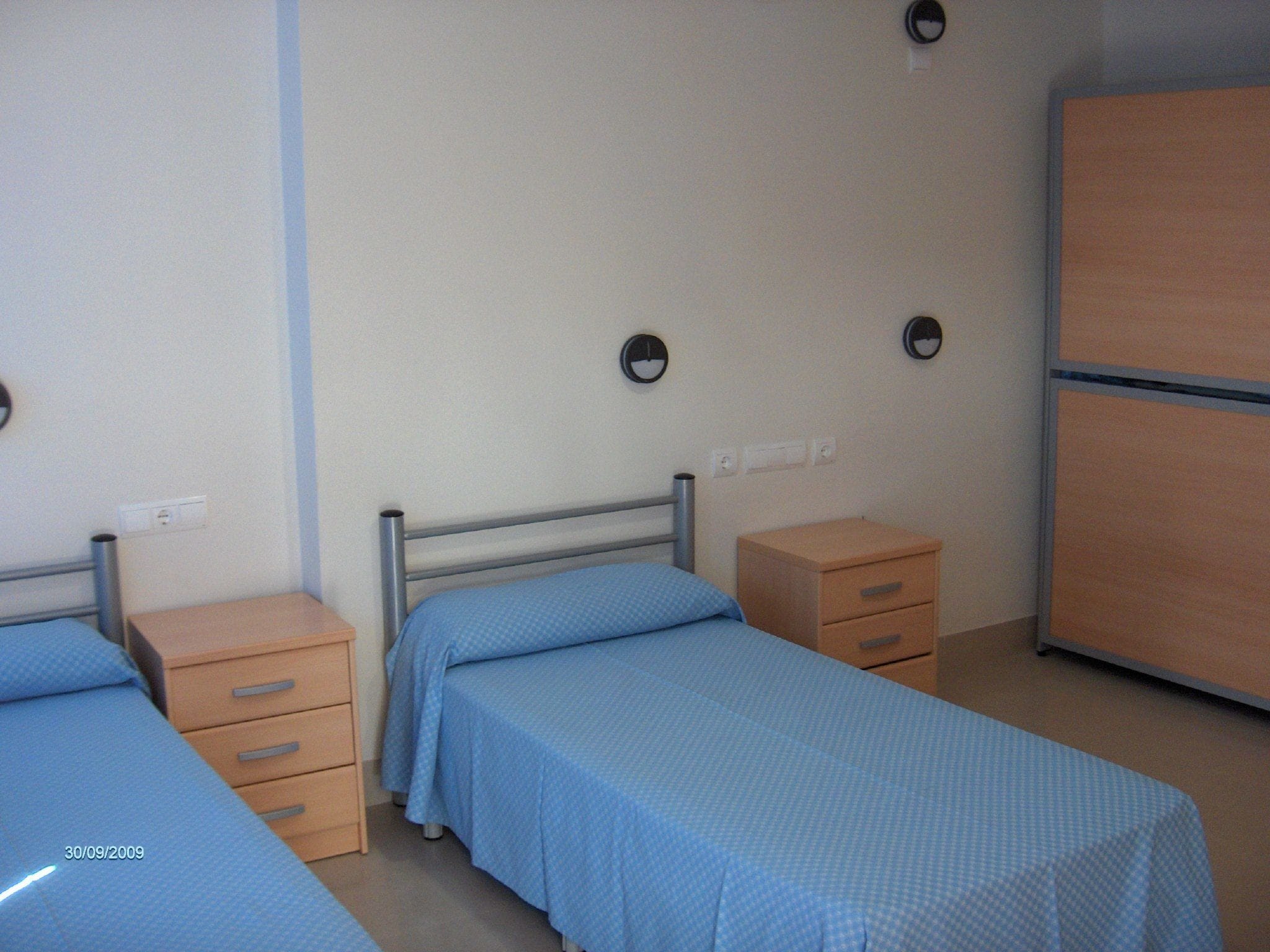 Una habitación con dos camas individuales, cada una con una mesita de noche de madera y un cajón. Se observa un armario contra la pared y dos relojes de pared.