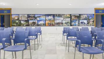 Een congreszaal met rijen blauwe stoelen, een betegelde vloer en een muur met verschillende foto's van stadsgezichten.