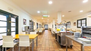Un'ampia mensa con lunghi tavoli e sedie, un'area cucina con elettrodomestici in acciaio inossidabile e un pavimento piastrellato.