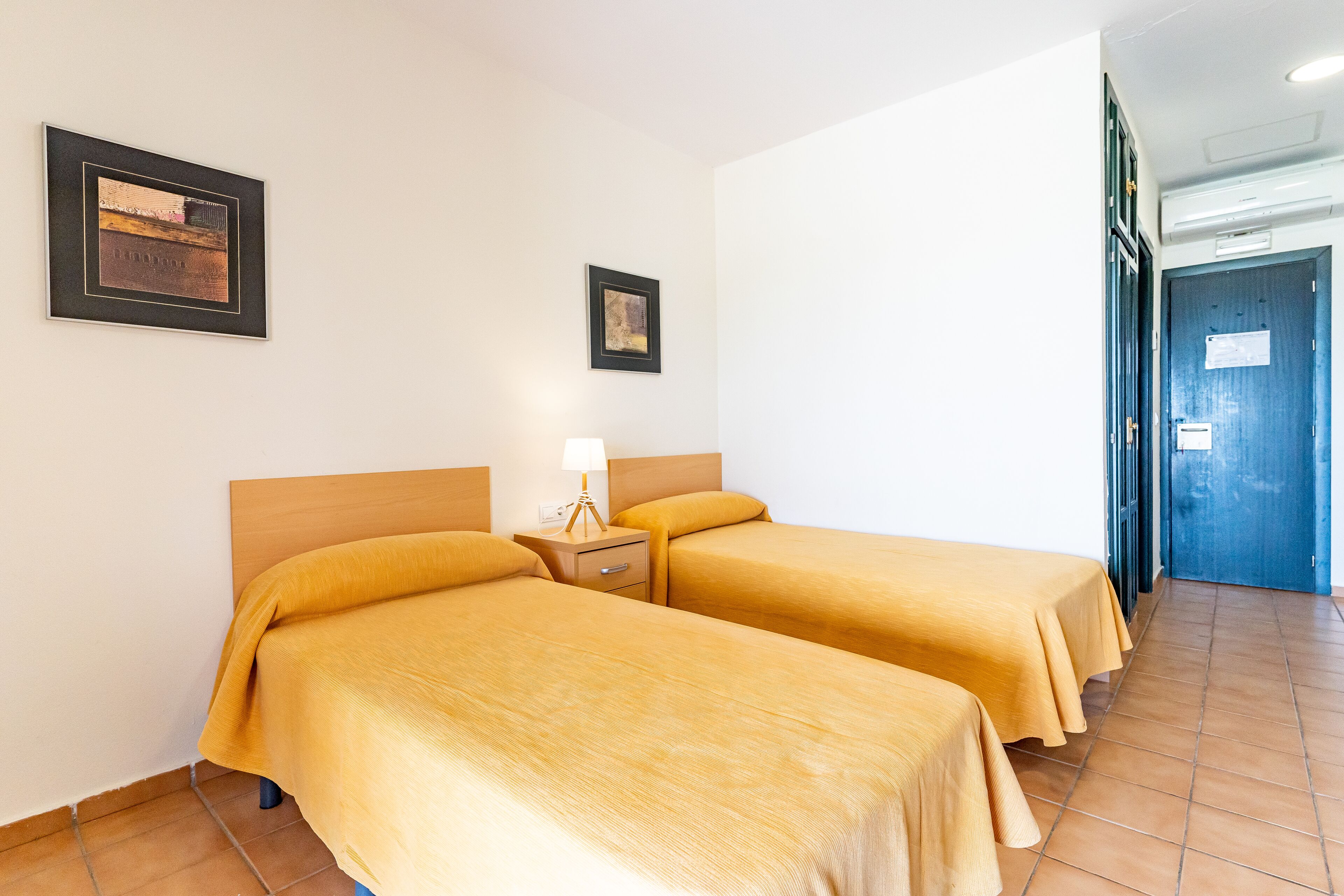 A hotel room with two beds, a nightstand with a lamp, and two framed pictures on the wall.