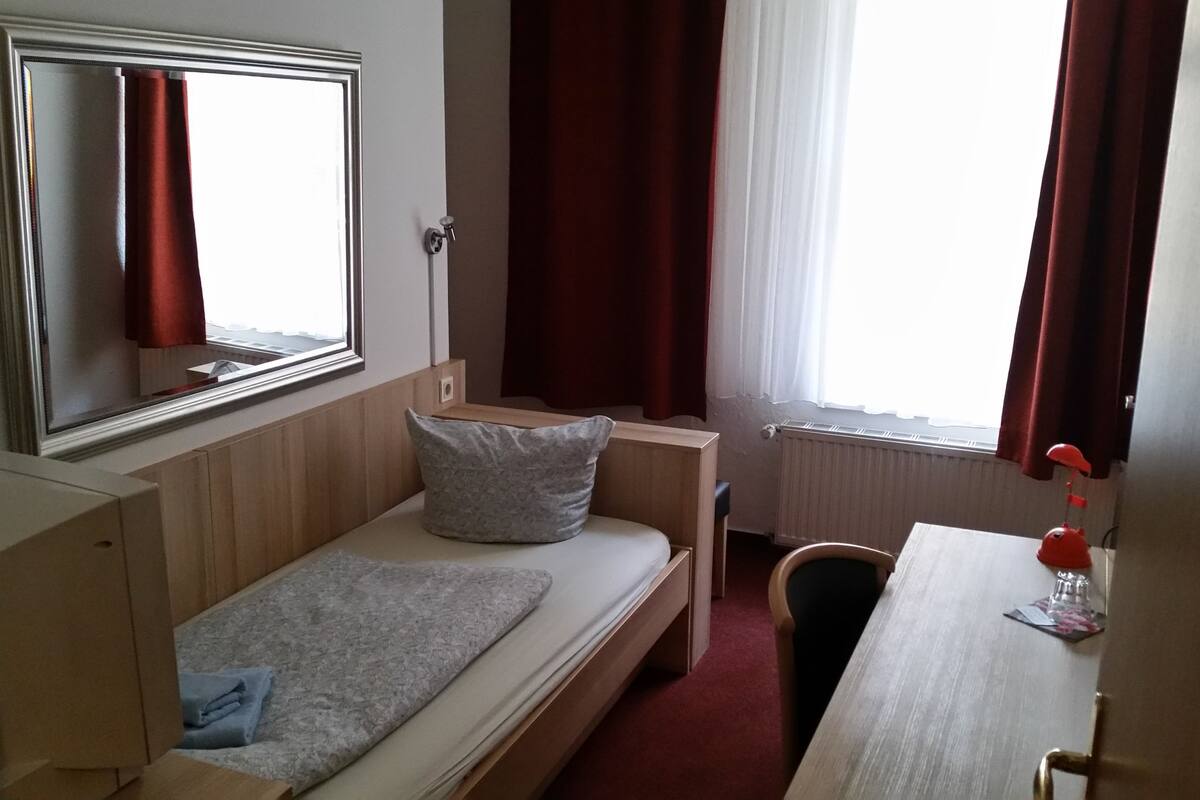 Una habitación pequeña con una cama individual, cabecera de madera, mesita de noche, escritorio y una silla. Cuenta con una ventana con cortinas, un radiador y un espejo en la pared.