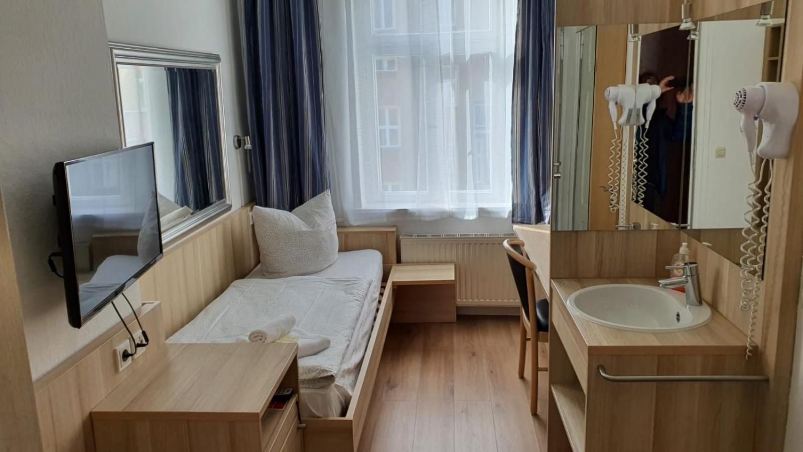 A compact hotel room with a bed, a TV, a bathroom with a sink and mirror, and a window with curtains.