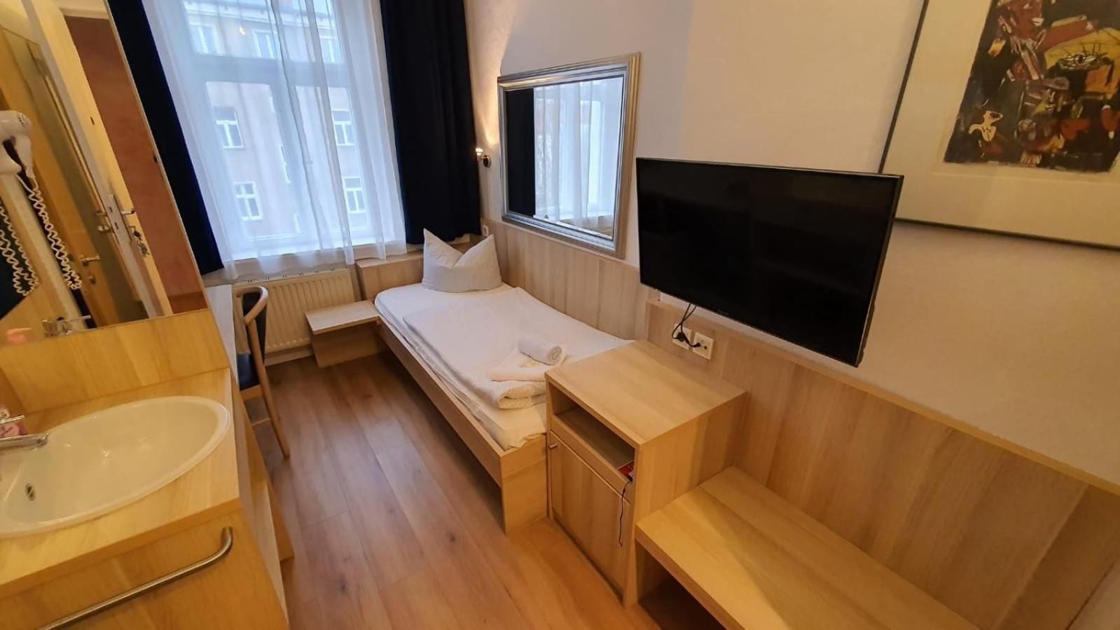 A compact room with a bed, a small bedside table, a built-in TV, a window with curtains, and a wooden floor.