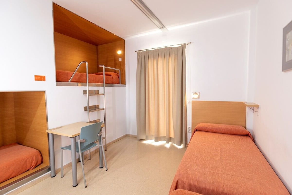 Een compacte hotelkamer met stapelbedden, een bureau, een stoel en een raam met gordijnen.