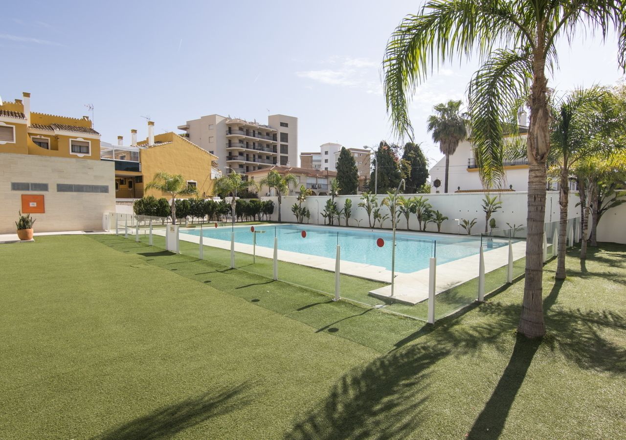 Une piscine rectangulaire entourée d’une clôture blanche, avec des palmiers et une pelouse au premier plan.