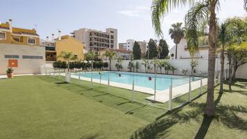 Une piscine rectangulaire entourée d’une clôture blanche, avec des palmiers et une pelouse au premier plan.