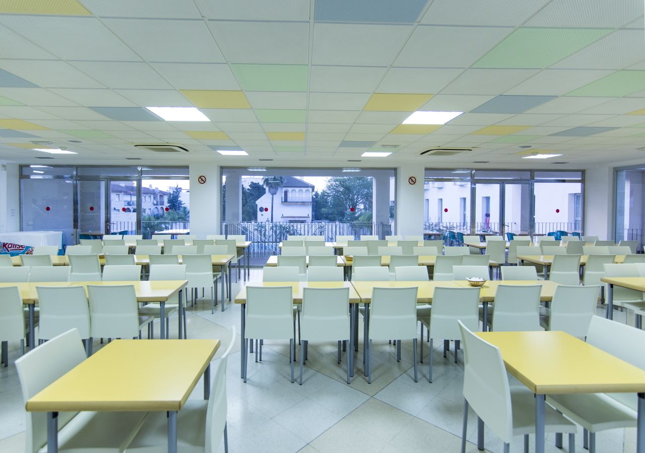 Een cafetaria met meerdere tafels en stoelen, grote ramen en uitzicht op gebouwen.