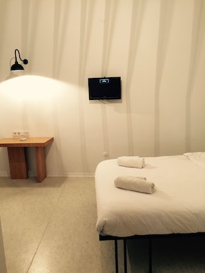 Una habitación de hotel con una cama, una mesita de noche de madera, un televisor montado en la pared y una lámpara.