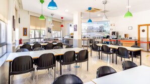 A cafeteria with multiple tables and chairs, a kitchen area in the background, and large windows allowing natural light. - Albergue Inturjoven Málaga - Hostel (Málaga)