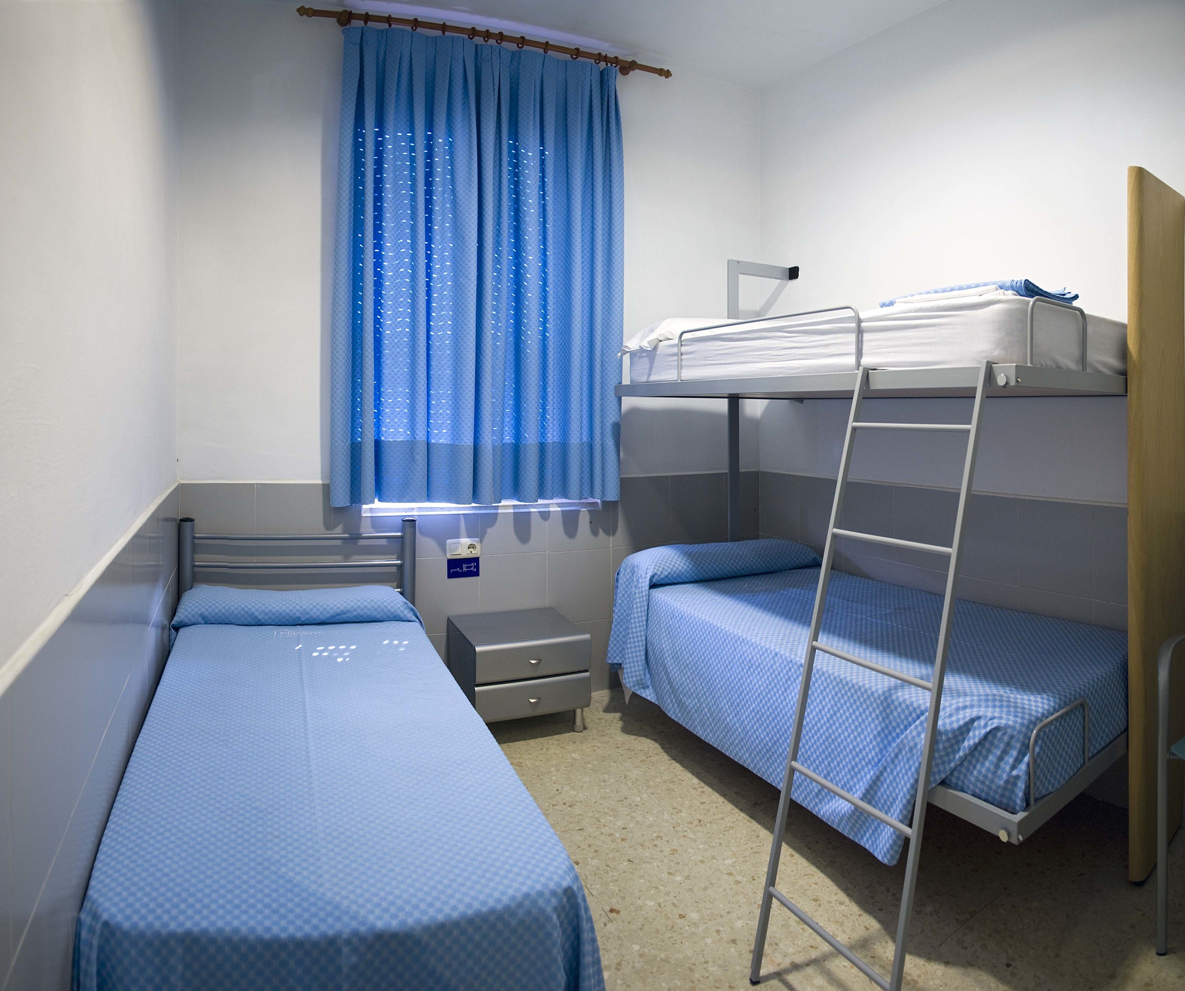 Un dormitorio con dos camas individuales, una litera, un ventanal con cortinas azules, un escritorio y una silla.