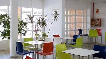 A spacious indoor dining area with multiple tables and chairs, large windows, and potted plants.