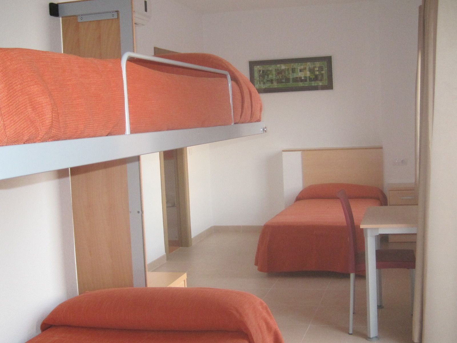 A dormitory room with bunk beds and a single bed, a small table, and a framed picture on the wall.
