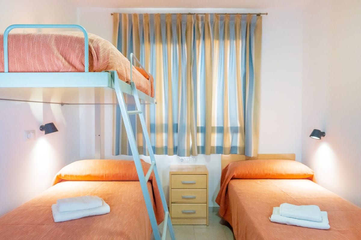 A room with two beds, one with a bunk bed and the other with a single bed, a small bedside table, and a window with curtains.
