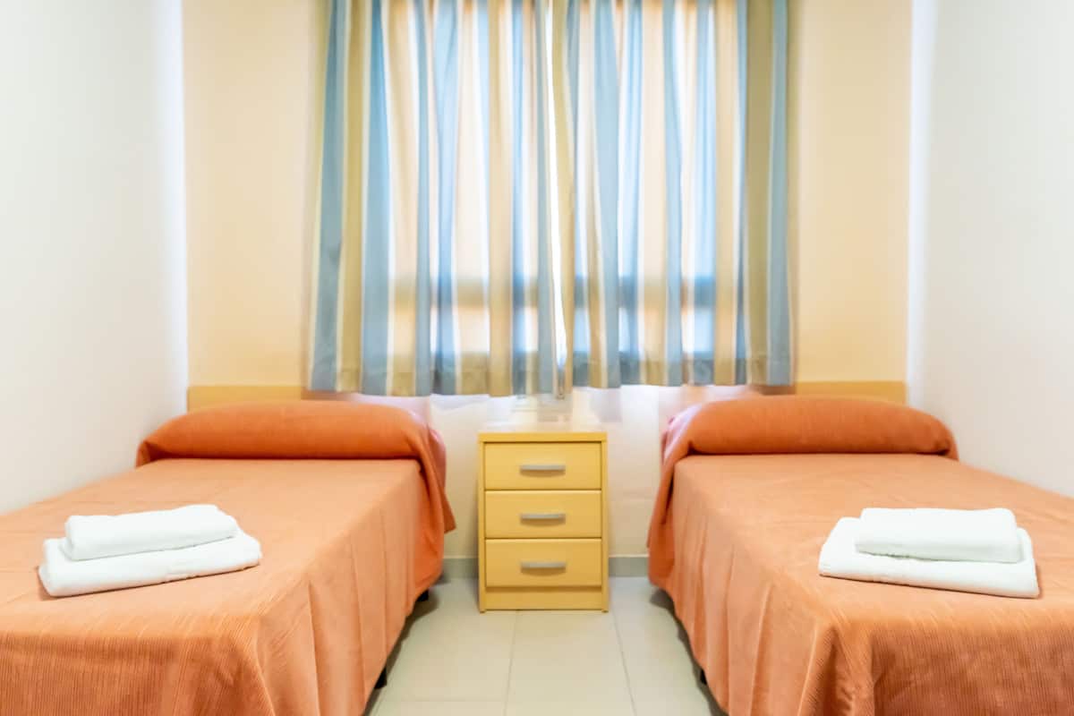 A hotel room with two beds, each with an orange bedspread and a white towel. There is a wooden nightstand between the beds with drawers.
