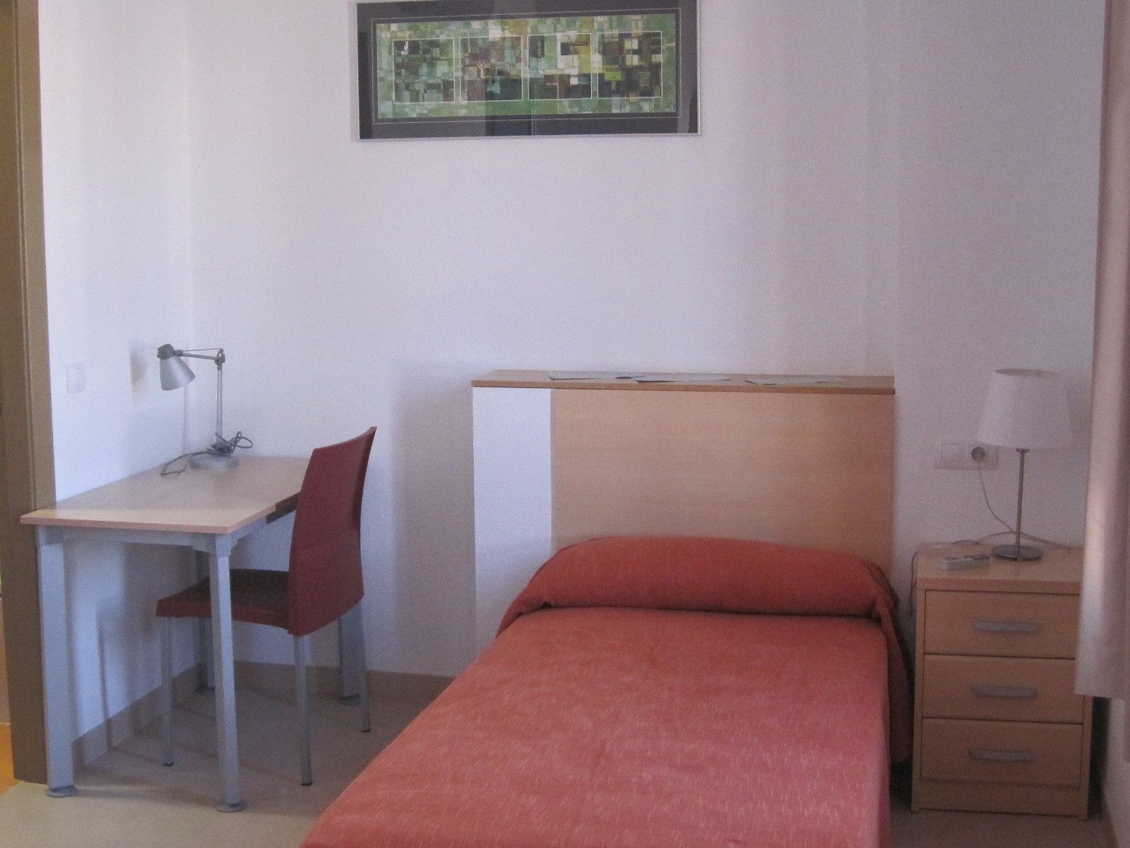 Una habitación pequeña con una cama, un escritorio, una silla y una mesita de noche.