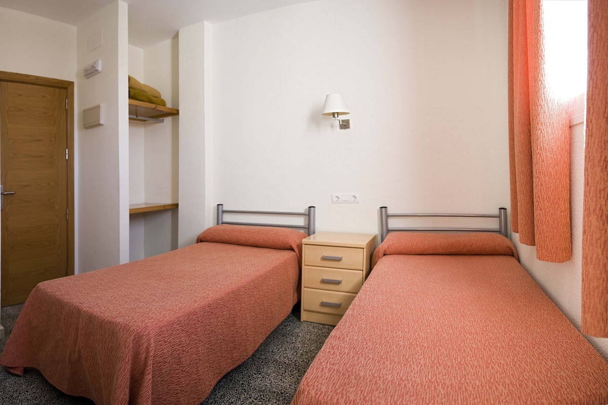 Una habitación pequeña y sencilla con dos camas individuales, una puerta de madera, un mueble bajo y una lámpara fijada a la pared.