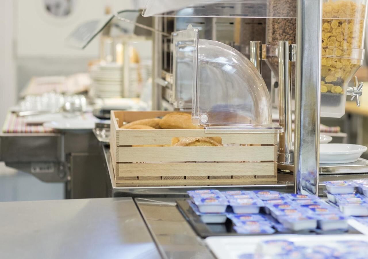 Disposição de buffet com uma cúpula de vidro transparente sobre uma caixa de madeira com doces, e diversos alimentos embalados no balcão.