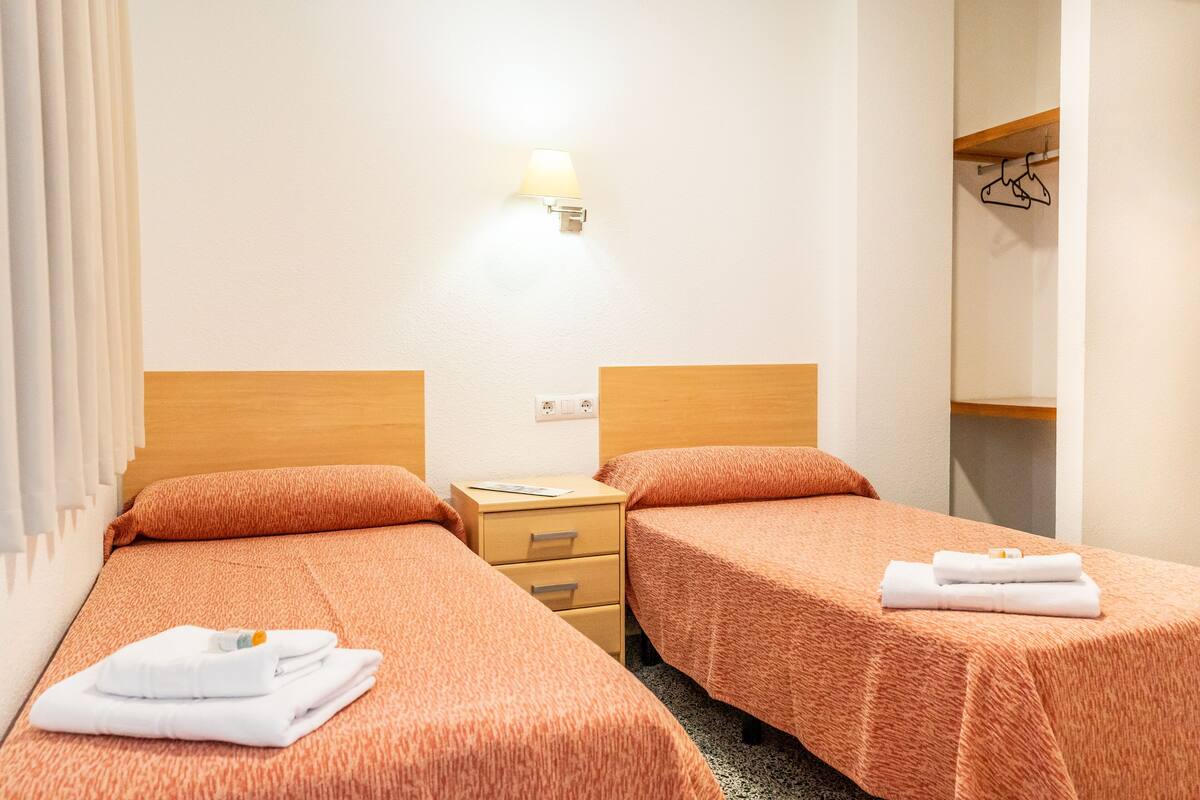 Habitación de hotel con dos camas individuales, una mesita de noche de madera, una lámpara de pared y un perchero.