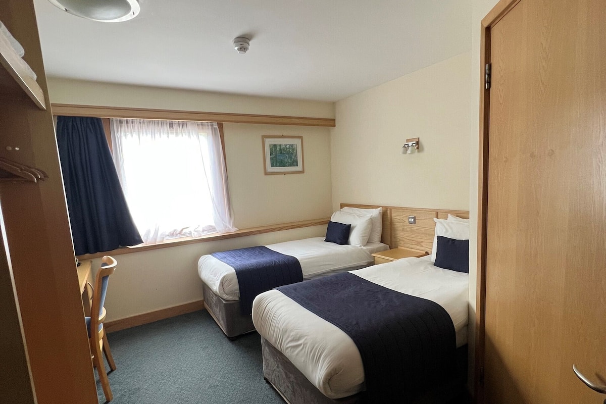 Una habitación de hotel con dos camas, una ventana con cortinas, un pequeño cuadro en la pared y una puerta.