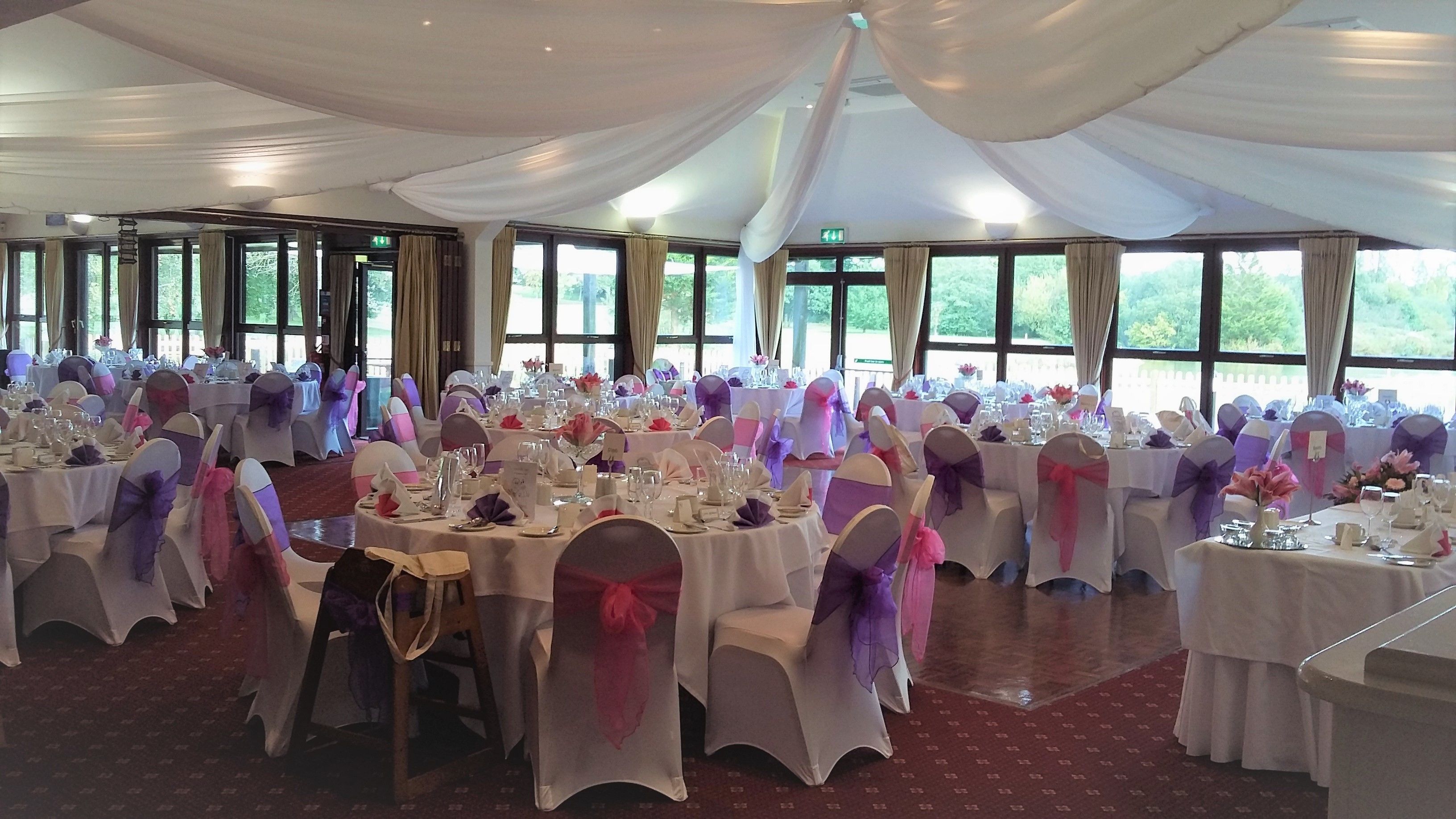 Un salón de banquetes con mesas redondas dispuestas para un evento formal, ventanas amplias y un techo alto adornado con telas.