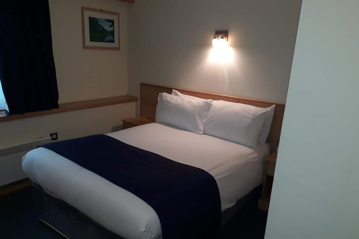 Habitación de hotel con cama, mesita de noche, luz empotrada en la pared y un cuadro enmarcado.
