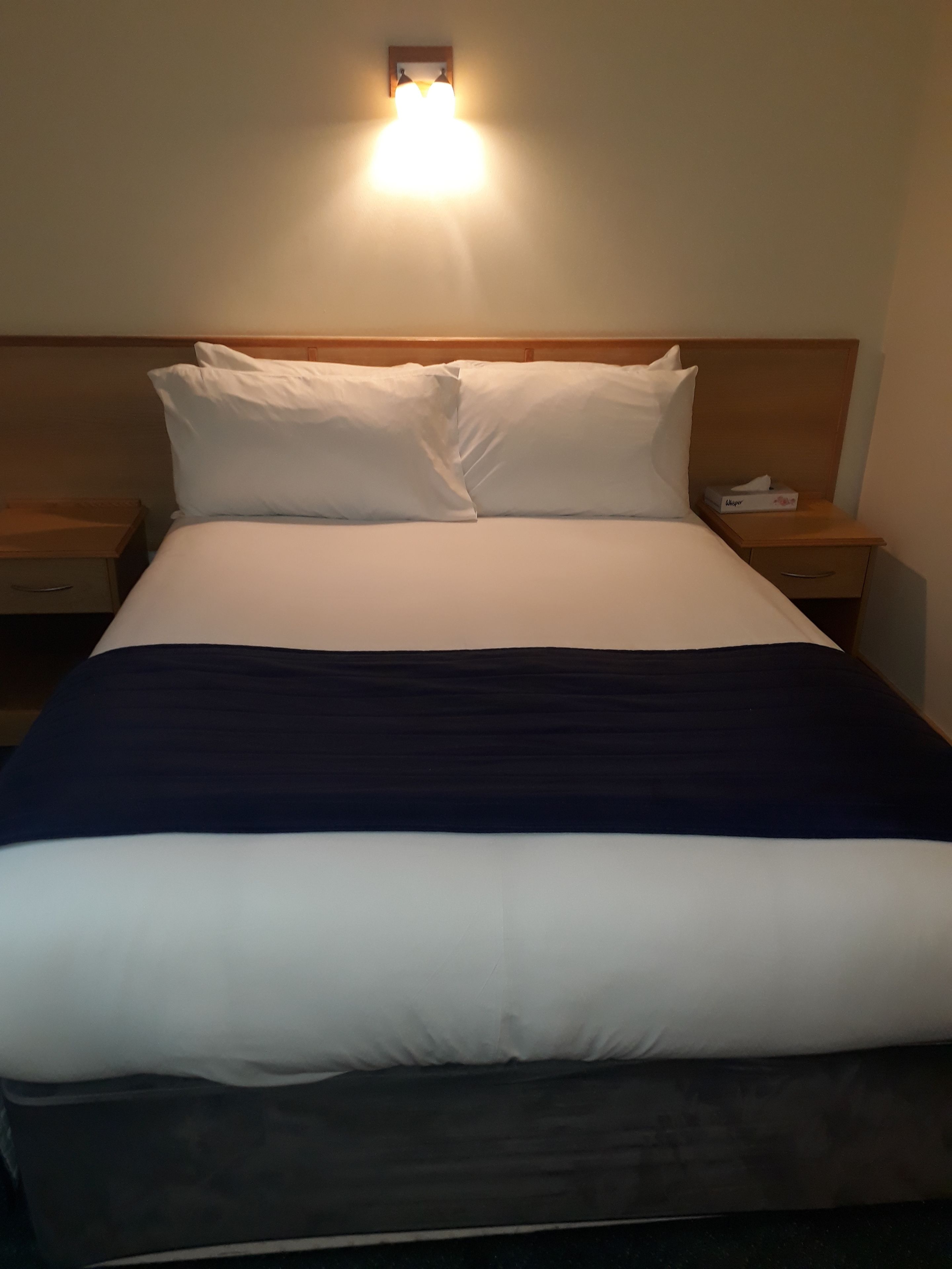 Un letto ben rifatto con biancheria nei toni del bianco e del blu navy, una testiera in legno e comodini illuminati da lampade.