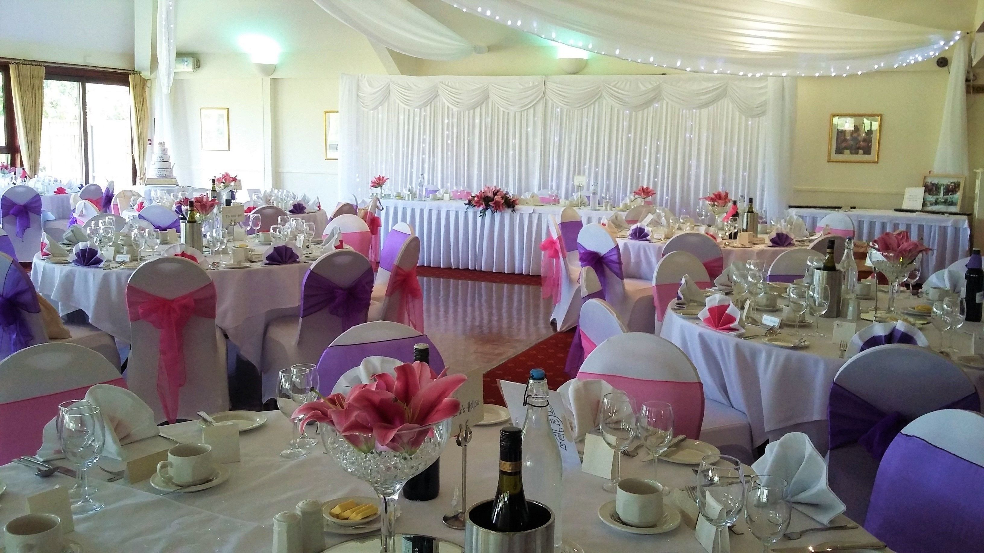 A banquet hall with round tables set for a formal event, featuring white tablecloths and purple and pink chair covers.