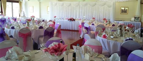 A banquet hall with round tables set for a formal event, featuring white tablecloths and purple and pink chair covers.