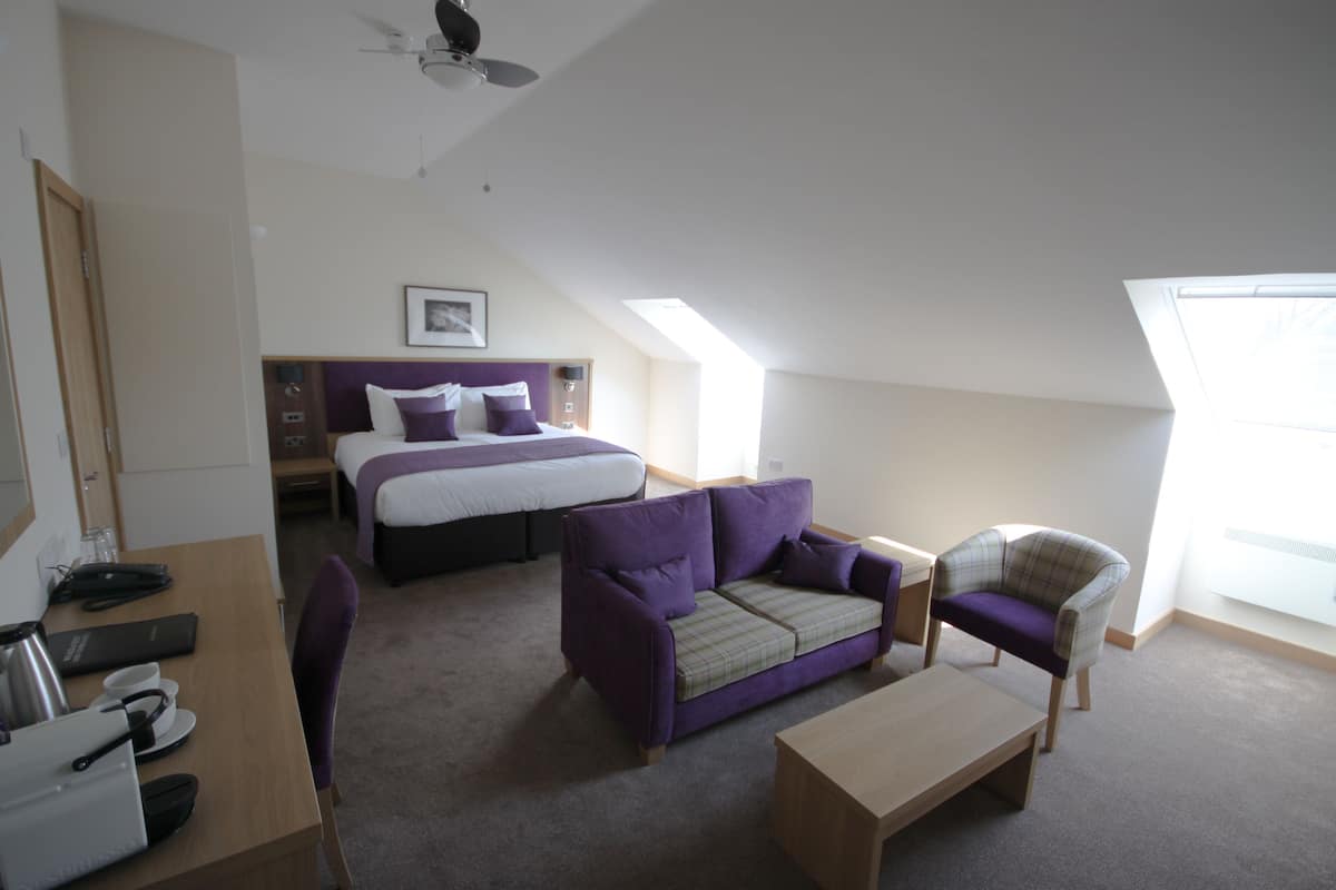 Una habitación de hotel con una cama, un sofá morado, un sofá a cuadros, una mesa de madera y un escritorio con una tetera y tazas.